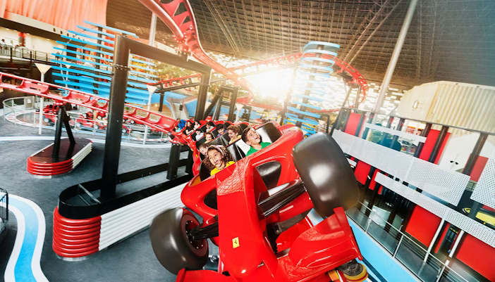 A sleek, red roller coaster with Ferrari branding, twisting through the air against the backdrop of Abu Dhabi's skyline at twilight. The coaster features multiple loops and corkscrews, with excited visitors queuing to ride.